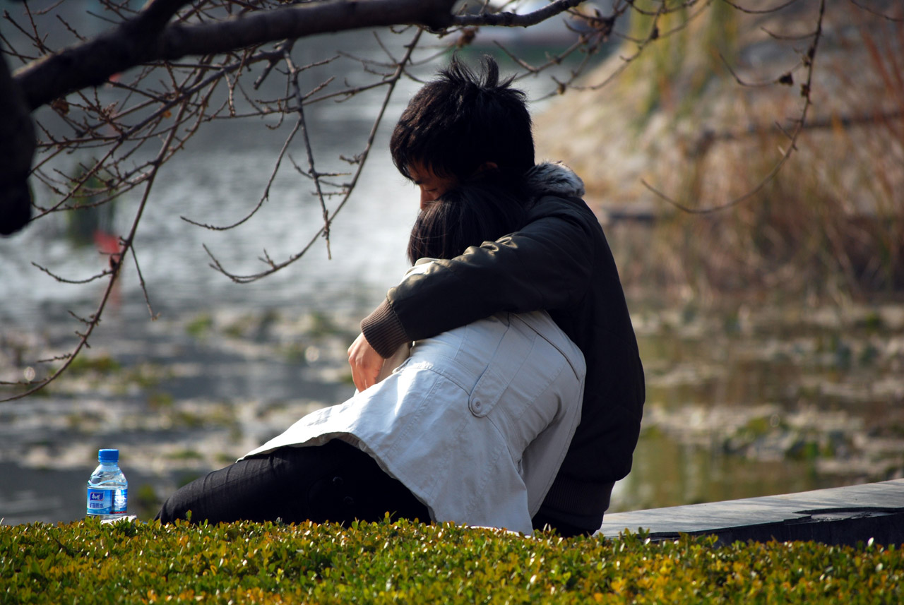 A young couple sit with their backs to the camera, facing a river, embracing with heads down.